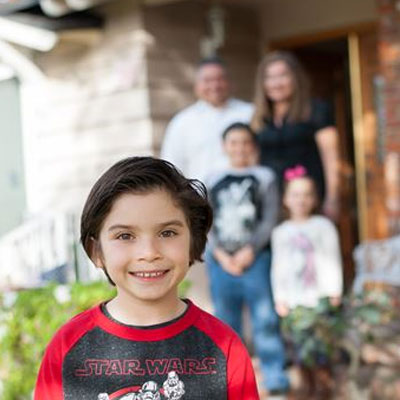 Emmanuel Peniche, standing outside his house with his family blurred in the background