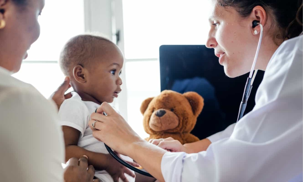 A physician using a stethoscope on a young patient while the mother holds his child's head up.