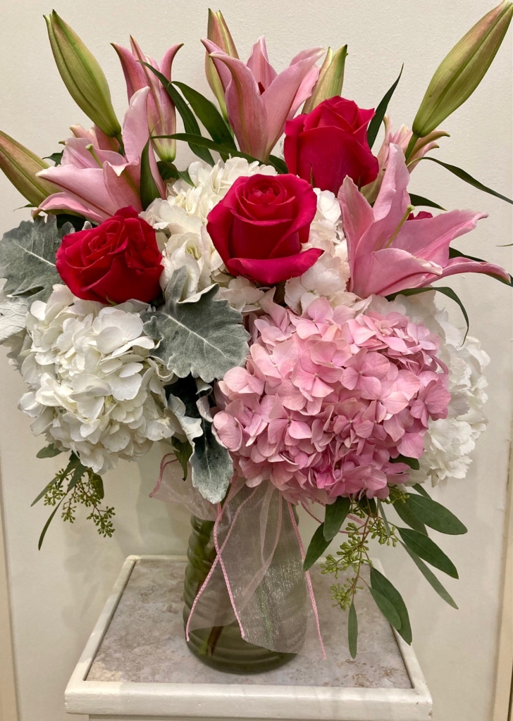 A bunch of red roses and white and pink flowers tied together with a pink ribbon in a clear vase