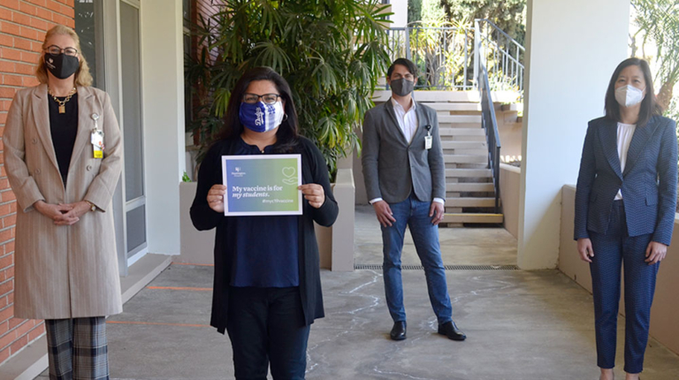 Masked employees spaced 6 feet apart with a banner saying vaccines for teachers