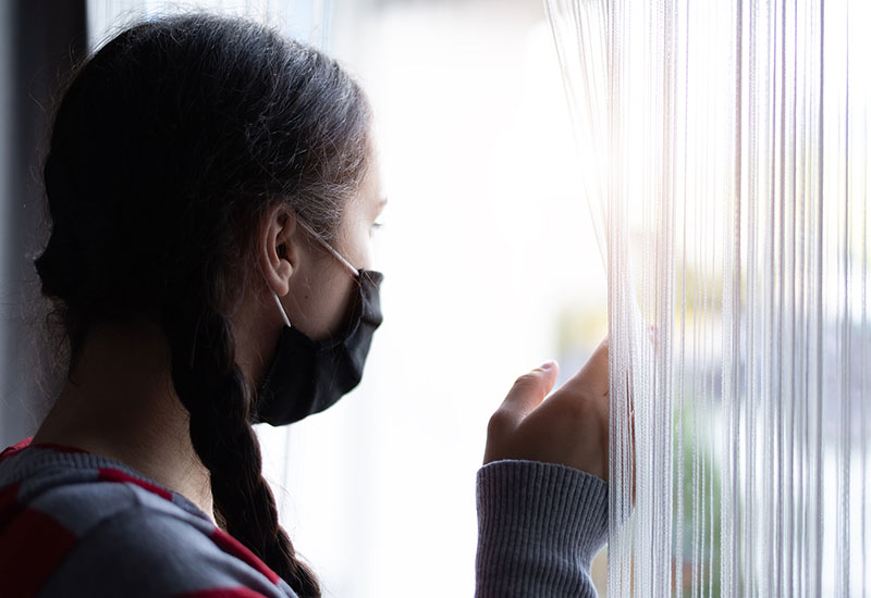 Teenager with mask on, looking outside through curtains