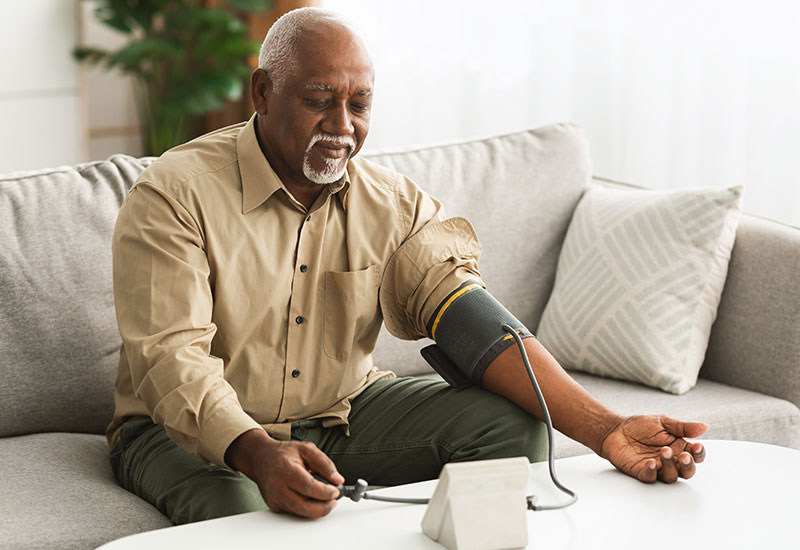 Senior man sitting on a couch and taking his own blood pressure with a small at-home machine