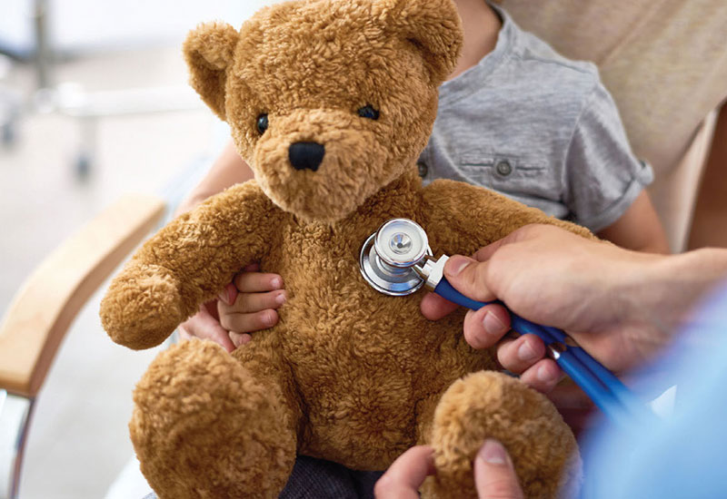 Child holding a stuffed teddy bear and doctor putting a stethoscope to the bear's chest