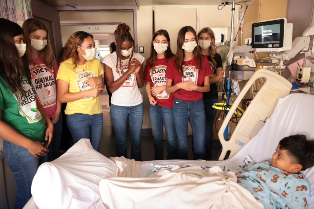 Masked volunteers in costume shirts visiting young child in hospital